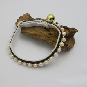 May include: A white cat collar with a black ribbon and a row of white pearl beads. A gold bell is attached to the collar.