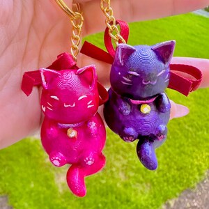 May include: Two cat-shaped keychains attached to gold-toned metal rings. One cat is bright pink and the other is dark purple, both featuring a textured finish, a small gold-colored bell at the neck, and a decorative red ribbon tied at the top.