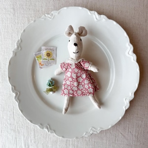 May include: A handmade white mouse doll wearing a pink floral dress with a lace collar. The doll is on a white scalloped plate, next to seed packets and a small floral decoration. The doll's ears are beige.