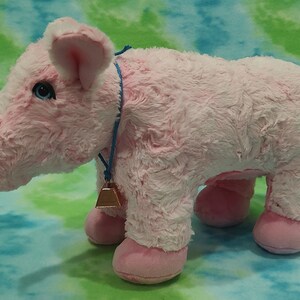 Pu&ograve; includere: Un giocattolo di maiale di peluche rosa con un cordoncino blu e un campanello dorato. Il maiale ha un naso rosa e zoccoli rosa.