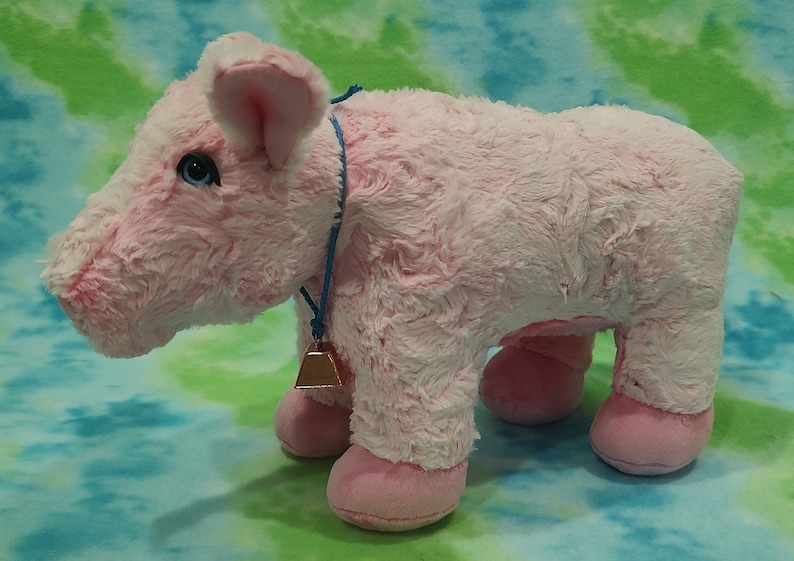Pu&ograve; includere: Un giocattolo di maiale di peluche rosa con un cordoncino blu e un campanello dorato. Il maiale ha un naso rosa e zoccoli rosa.
