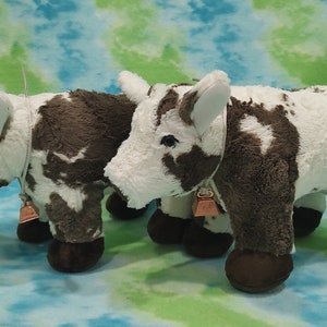 Pu&ograve; includere: Due giocattoli di peluche a forma di mucca, marroni e bianchi, con campane intorno al collo. Le mucche sono in piedi una accanto all'altra su uno sfondo blu e verde.