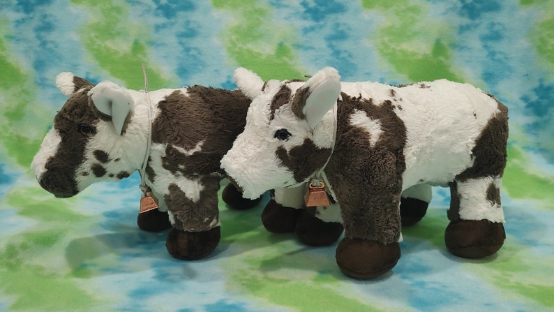 Pu&ograve; includere: Due giocattoli di peluche a forma di mucca, marroni e bianchi, con campane intorno al collo. Le mucche sono in piedi una accanto all'altra su uno sfondo blu e verde.