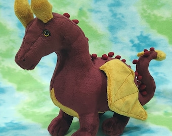 May include: A plush burgundy dragon toy with yellow wings and red spots. The dragon has a long tail and large horns.