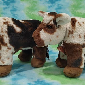 Pu&ograve; includere: Due figurine di mucca di peluche marroni e bianche macchiate con campane intorno al collo. Le mucche sono in piedi una accanto all'altra su uno sfondo blu e verde.