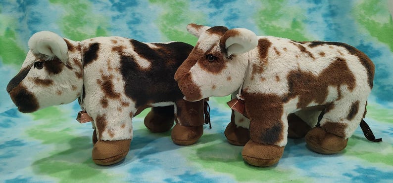Pu&ograve; includere: Due figurine di mucca di peluche marroni e bianche macchiate con campane intorno al collo. Le mucche sono in piedi una accanto all'altra su uno sfondo blu e verde.