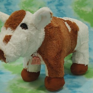 Pu&ograve; includere: Una mucca di peluche marrone e bianca con una campana intorno al collo. La mucca &egrave; in piedi su uno sfondo blu e verde.
