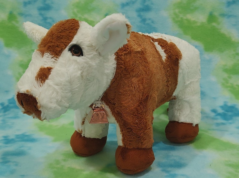 Pu&ograve; includere: Una mucca di peluche marrone e bianca con una campana intorno al collo. La mucca &egrave; in piedi su uno sfondo blu e verde.