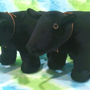 Pu&ograve; includere: Due giocattoli di peluche di toro neri con cinturini in pelle marrone intorno al collo. I giocattoli hanno occhi neri e sono in piedi su uno sfondo blu e verde.