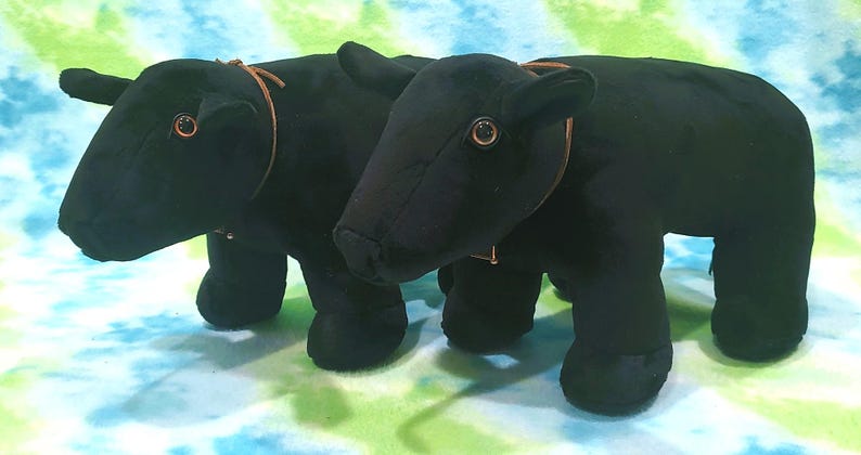 Pu&ograve; includere: Due giocattoli di peluche di toro neri con cinturini in pelle marrone intorno al collo. I giocattoli hanno occhi neri e sono in piedi su uno sfondo blu e verde.