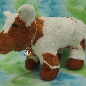 Pu&ograve; includere: Una mucca di peluche marrone e bianca con un campanello intorno al collo. La mucca &egrave; in piedi su uno sfondo blu e verde.