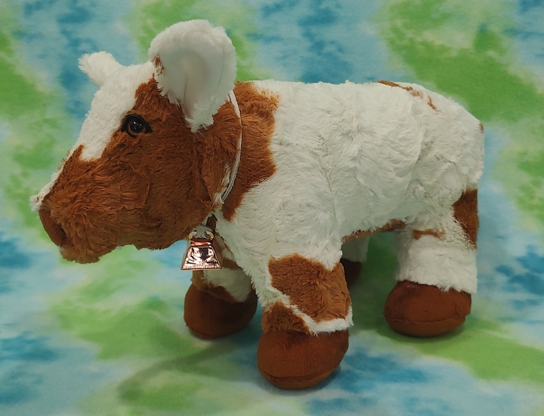 Pu&ograve; includere: Una mucca di peluche marrone e bianca con un campanello intorno al collo. La mucca &egrave; in piedi su uno sfondo blu e verde.