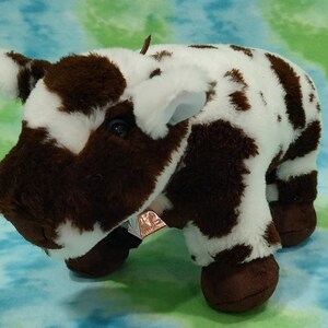 Pu&ograve; includere: Una mucca di peluche marrone e bianca con il naso nero e gli zoccoli marroni. La mucca &egrave; sdraiata con la testa girata di lato.