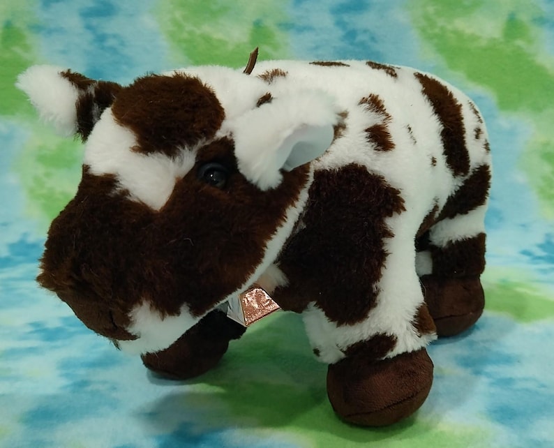 Pu&ograve; includere: Una mucca di peluche marrone e bianca con il naso nero e gli zoccoli marroni. La mucca &egrave; sdraiata con la testa girata di lato.