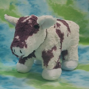 Pu&ograve; includere: Un giocattolo di peluche di mucca bianco e viola con il naso marrone e gli occhi neri. La mucca &egrave; in piedi su uno sfondo blu e verde.