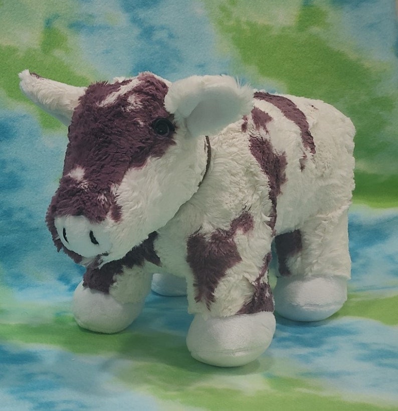 Pu&ograve; includere: Un giocattolo di peluche di mucca bianco e viola con il naso marrone e gli occhi neri. La mucca &egrave; in piedi su uno sfondo blu e verde.