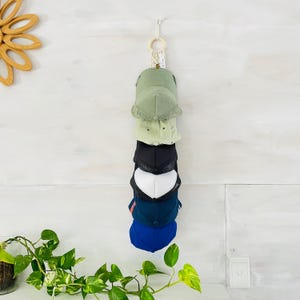 May include: A wall-mounted hat organiser displaying six baseball caps in various colours: olive green, light green, black, white, navy, and royal blue. The organiser is suspended from a macrame loop and hangs against a white wall.