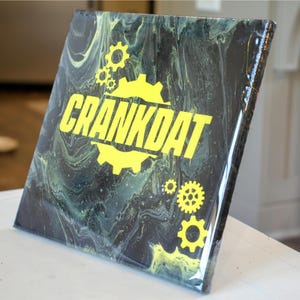 May include: Square canvas art with a black and green marbled background. The word "CRANKDAT" is in bold yellow letters, with yellow gear graphics. The artwork is displayed on a white surface.