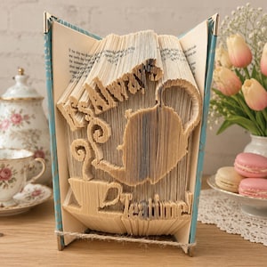 May include: A folded book art sculpture featuring the text Always Tea Time with a teapot and teacup silhouette. The pages are intricately creased to form the shapes and words, with a light blue book cover visible on the sides of the open book.