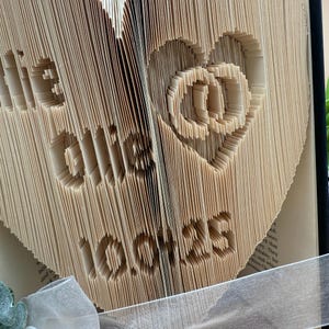 May include: A book art sculpture in the shape of a heart with the words "Be Kind" and "Love" carved into the pages. The book is made of beige paper and is wrapped in a white ribbon.