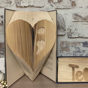 Può includere: Un libro piegato a forma di cuore con un punto e virgola all'interno. Il libro è appoggiato su una superficie di legno davanti a un muro di mattoni.
