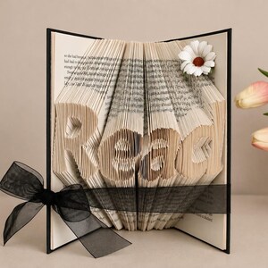 May include: A book with folded pages forming the word Read, featuring a black ribbon tied around the center and a white daisy flower accent on the top right corner. The book has a black cover and contains printed text on the interior pages.