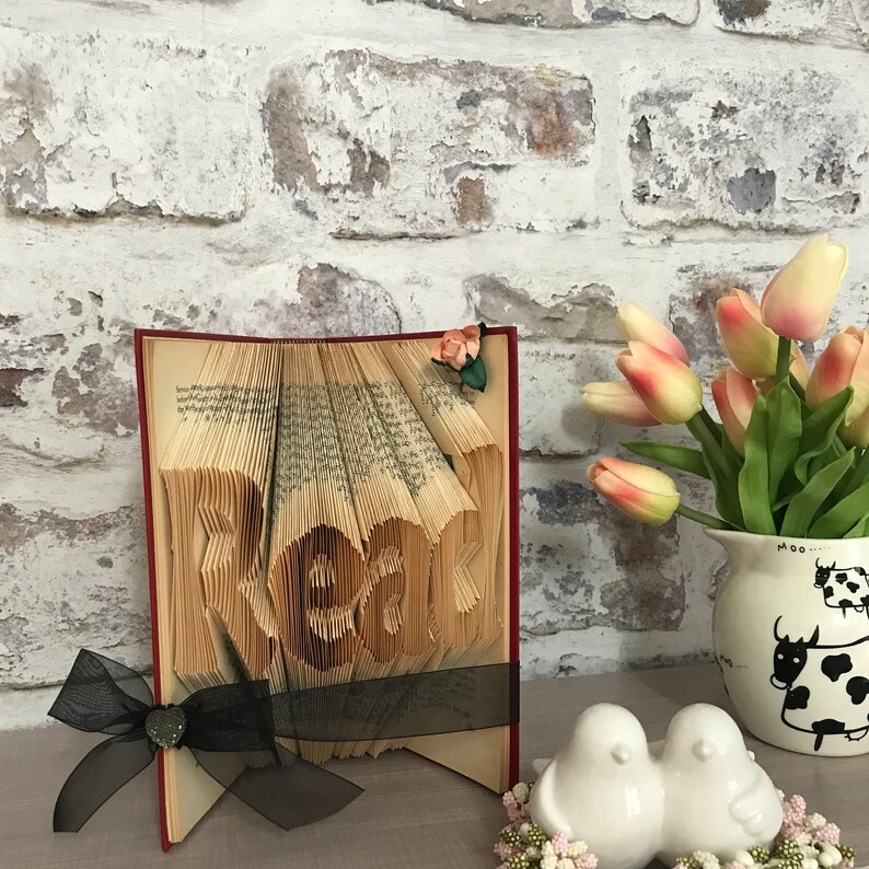 May include: A folded book art sculpture with the word Read formed by the pages. The book features a black ribbon tied around the cover with a small heart charm and a decorative fabric flower. Beside it sits a white pitcher with cow illustrations.