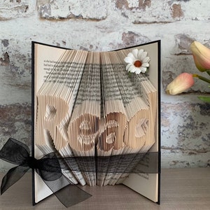May include: A folded book art sculpture with pages intricately creased to form the word "Read" in a central block. The book is accented with a white daisy flower on the top right corner and a sheer black ribbon tied around the base of the pages.