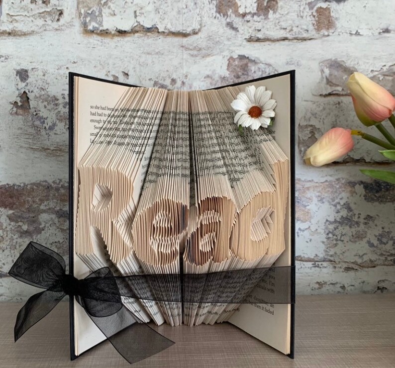 May include: A folded book art sculpture with pages intricately creased to form the word "Read" in a central block. The book is accented with a white daisy flower on the top right corner and a sheer black ribbon tied around the base of the pages.