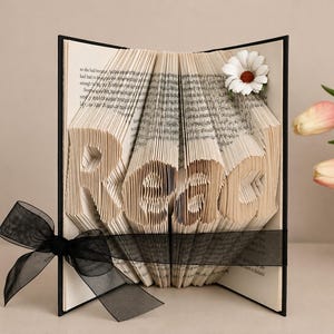 May include: A book with folded pages forming the word Read, featuring a black ribbon tied around the center and a white daisy flower accent on the top right corner. The book has a black cover and contains printed text on the interior pages.