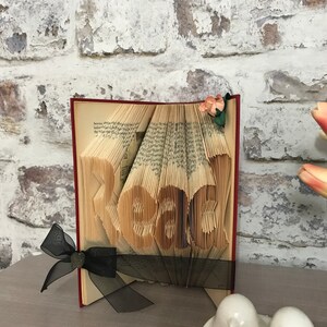 May include: A folded book art sculpture with pages intricately creased to form the word Read. The book features a dark red cover, a black sheer ribbon tied around the base, and a small artificial peach-colored flower attached to the top corner.
