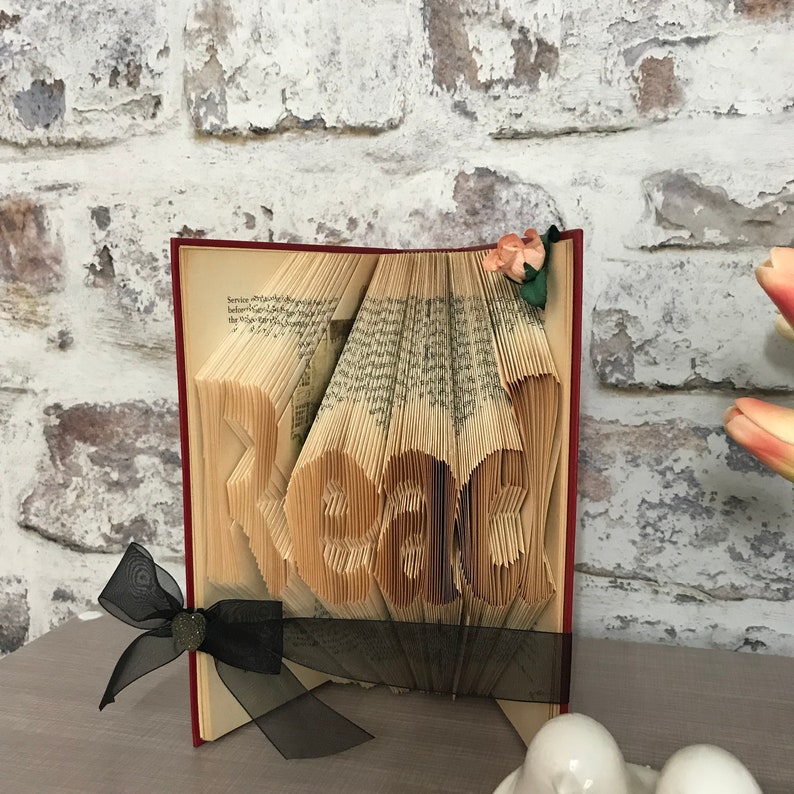 May include: A folded book art sculpture with pages intricately creased to form the word Read. The book features a dark red cover, a black sheer ribbon tied around the base, and a small artificial peach-colored flower attached to the top corner.