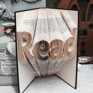 May include: A book sculpture with pages folded to form the word Read in a clear, legible font. The book is open with a black cover, and the paper edges are visible, creating a three-dimensional effect where the letters stand out from the pages.