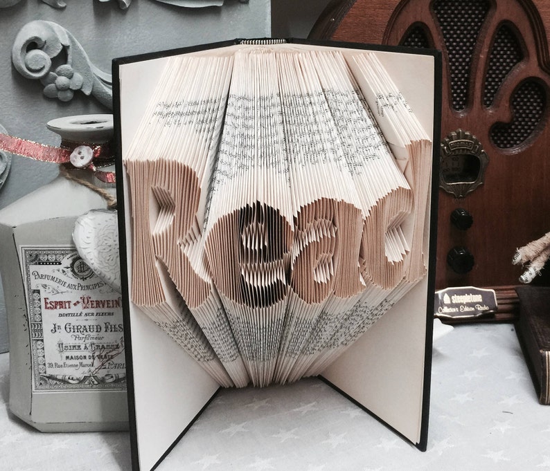 May include: A book sculpture with pages folded to form the word Read in a clear, legible font. The book is open with a black cover, and the paper edges are visible, creating a three-dimensional effect where the letters stand out from the pages.