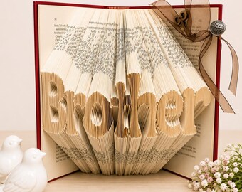 May include: A folded book art sculpture with pages intricately creased to form the word Brother in a bold font. The book features a deep red cover and is decorated with a sheer brown ribbon, a small metal key charm, and a circular silver button.