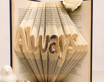 May include: A book with pages folded to form the word Always... in a vertical orientation. The book has a dark blue cover and is decorated with a single cream-colored artificial rose at the top right corner and a small white bird figurine.