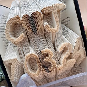 May include: A book art sculpture in the shape of a heart and the numbers 10324. The sculpture is made from the pages of a book that have been carefully folded and cut to create the design.