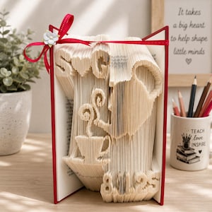 May include: A folded book art sculpture featuring the letters S and G separated by a heart, a teapot pouring into a cup, and the numbers 8.1.25. The book has a red cover and is decorated with a red ribbon tied in a bow with a small white flower.