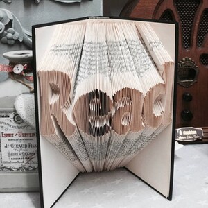 May include: A book with pages folded to form the word Read in a three-dimensional style. The book has a black cover and white pages with black printed text. The folded pages create a sculptural effect, standing upright with the word clearly visible.