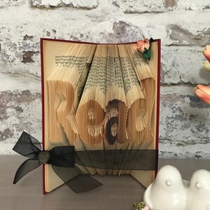 May include: A folded book art sculpture with pages intricately creased to form the word Read. The book features a dark red spine, a black sheer ribbon tied with a small heart-shaped charm, and a small artificial peach-colored flower on the top corner.