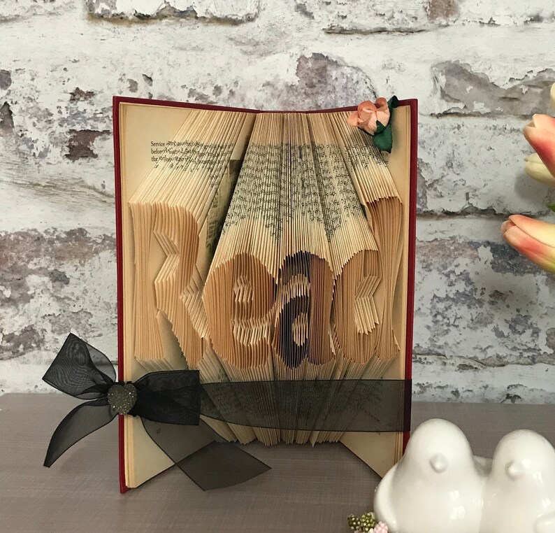 May include: A folded book art sculpture with pages intricately creased to form the word Read. The book features a dark red spine, a black sheer ribbon tied with a small heart-shaped charm, and a small artificial peach-colored flower on the top corner.
