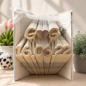 May include: A folded book art sculpture featuring the letters G and G separated by a heart shape, with the date 16.06.22 displayed below. The pages are intricately creased to form these characters, and a white ribbon is tied around the top.