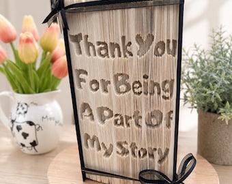 May include: A folded book art sculpture featuring the text Thank You For Being A Part Of My Story. The pages are intricately folded to create the lettering, and the book is accented with black ribbon bows at the top left and bottom right corners.