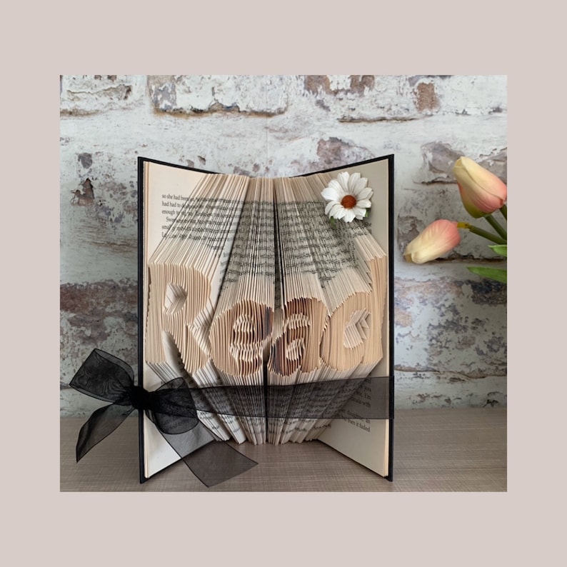 May include: A book art sculpture with pages folded to form the word Read. The book is accented with a black sheer ribbon tied around the center and a single white daisy flower attached to the top right corner of the pages, creating a decorative display.