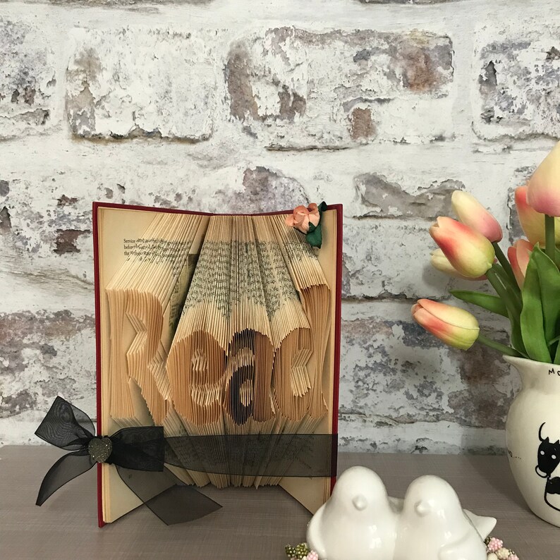 May include: A book art sculpture with pages folded to form the word Read. The book has a red cover and is decorated with a black sheer ribbon tied in a bow at the bottom left and a small artificial flower at the top right corner of the pages.