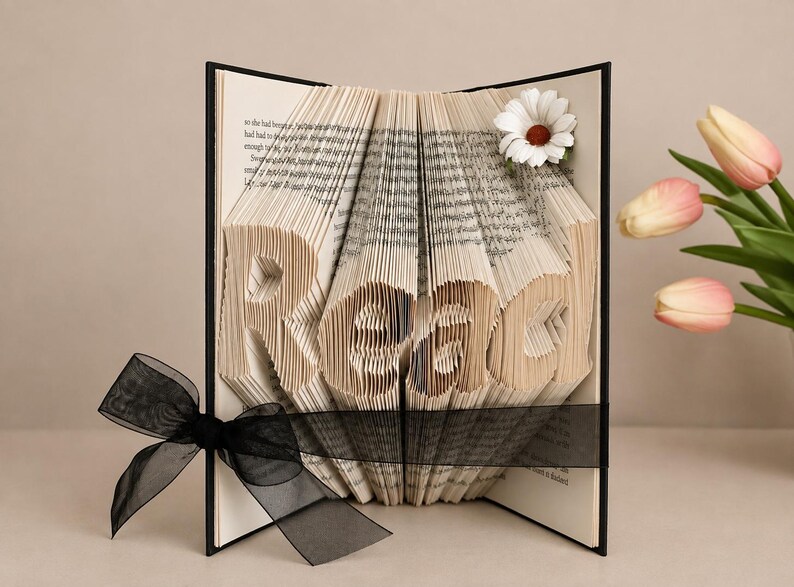 May include: A book with folded pages forming the word Read, featuring a black ribbon tied around the center and a white daisy flower accent on the top right corner. The book has a black cover and contains printed text on the interior pages.