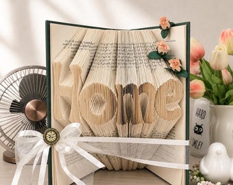 May include: A folded book art sculpture displaying the word home, featuring pages intricately creased to form the letters. The book is adorned with a white sheer ribbon, a small metallic circular embellishment, and three small peach-colored fabric roses.