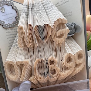 May include: A book art sculpture in the shape of the words "Love 07 08" with a grey flower on the side. The sculpture is made from the pages of a book that have been folded and cut to create the design.