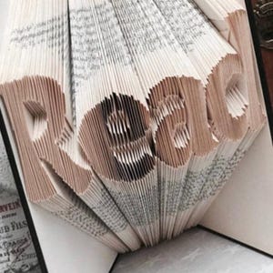 May include: A folded book art sculpture featuring the word Read created by precisely folding the individual pages of a hardcover book. The cream-colored paper edges are arranged to form the letters R, e, a, and d in a vertical orientation.