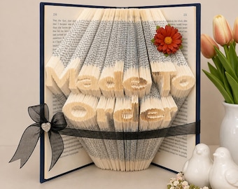 May include: A book with pages folded to form the text Made To Order, featuring a dark blue cover, a black sheer ribbon with a small heart-shaped rhinestone accent, and a single dark orange flower attached to the top right corner of the pages.